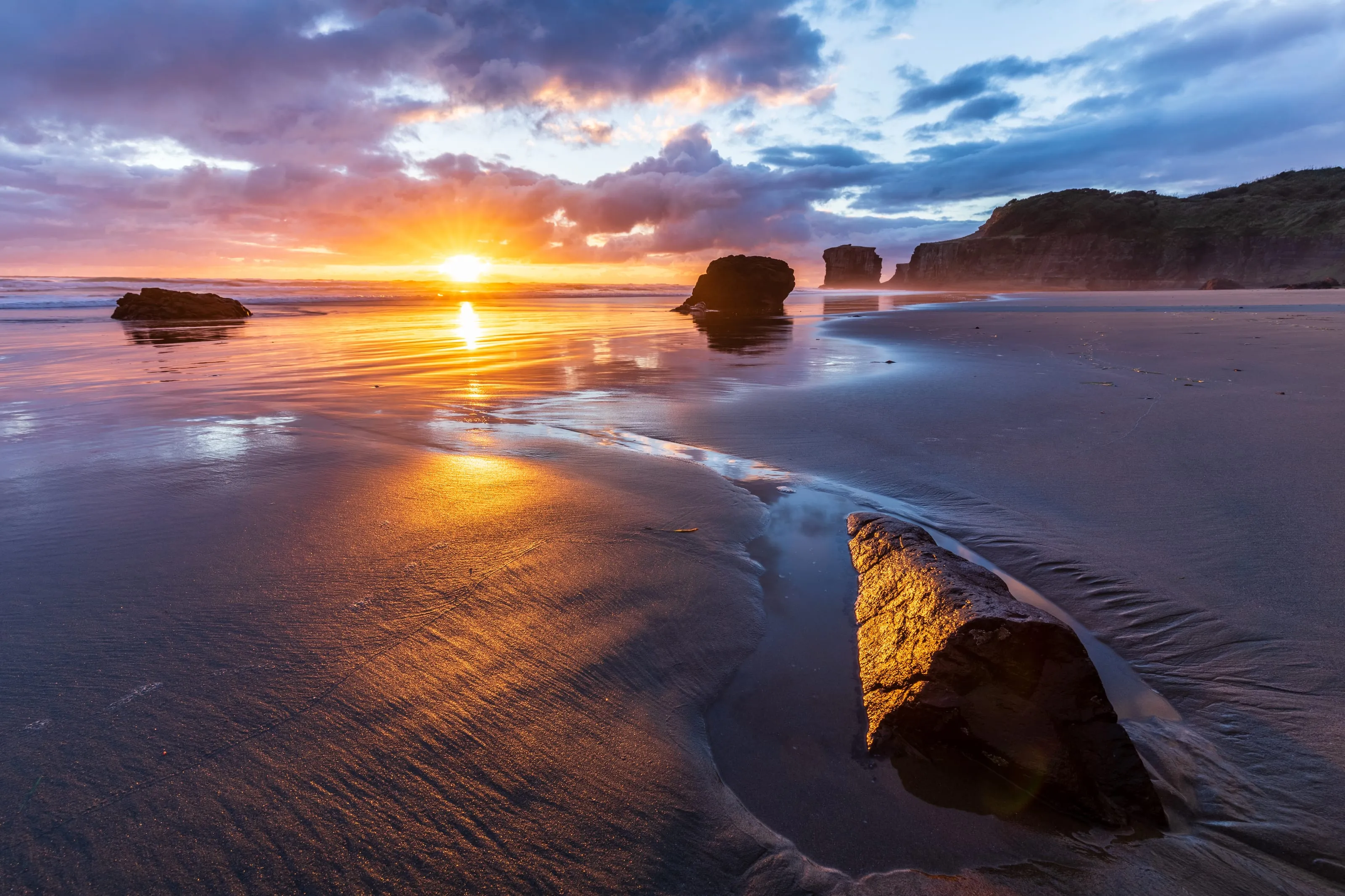 Sunset over Maori Bay, the sun low on the horizon behind sea stacks with the light running up the wet sand